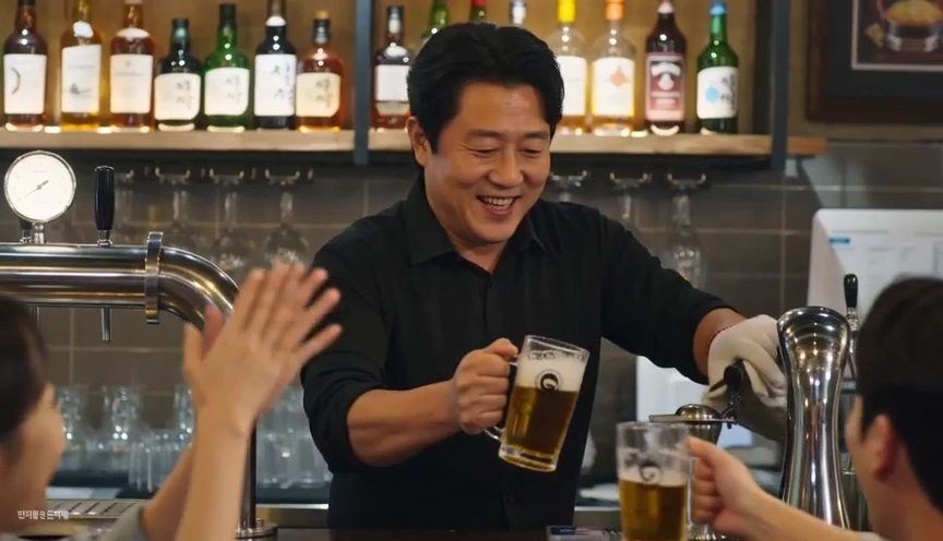 한국 광고 장면: 펍에서 점주가 웃으며 맥주를 건네는 모습